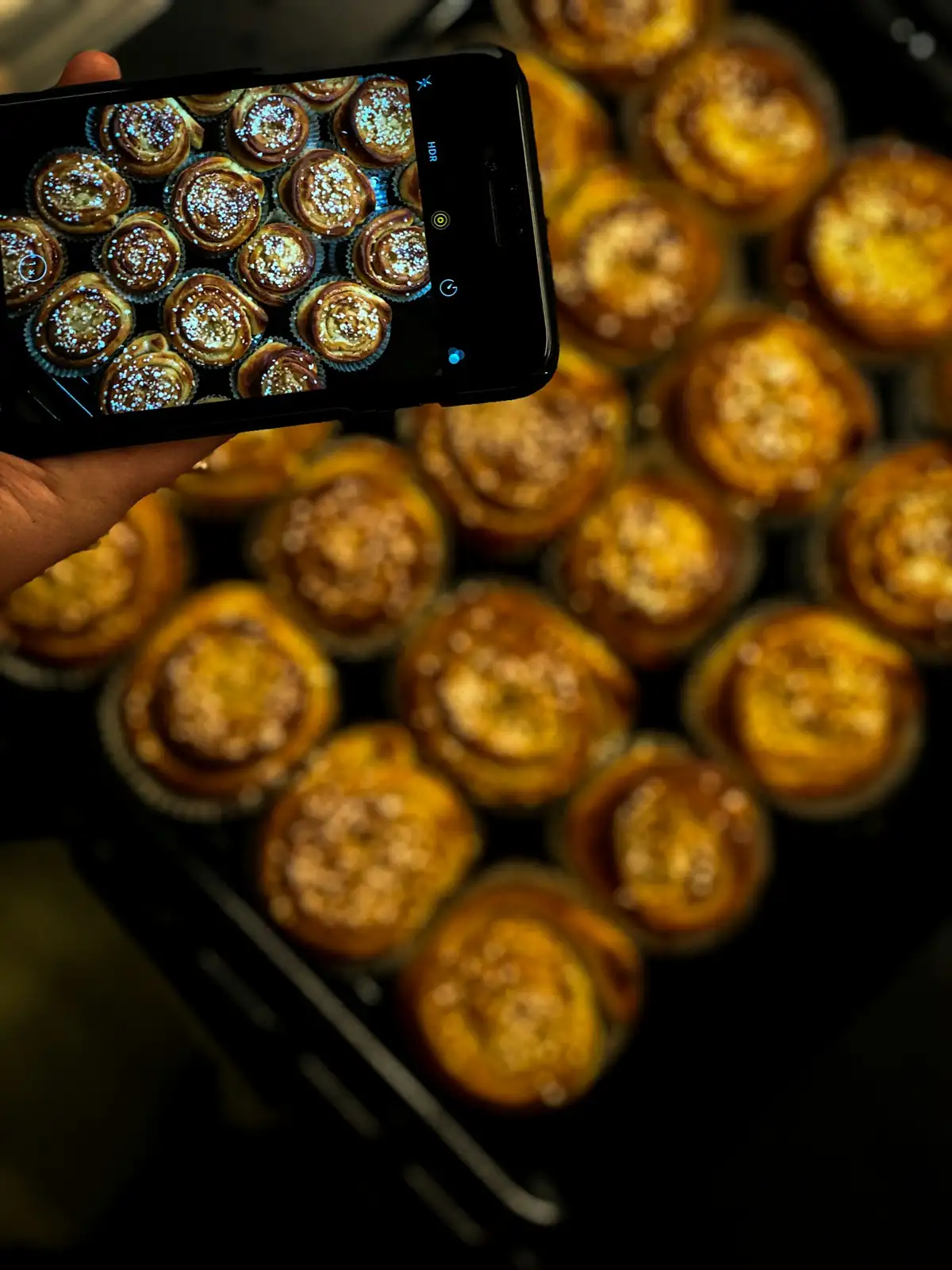 Mão segurando um celular que fotografa uma assadeira de pãezinhos de canela dourados; a tela mostra a mesma cena e os pães ao fundo estão fora de foco.